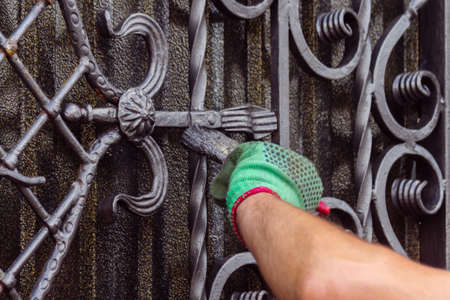 A man paints a gate with forged elements. On his hand is a green glove, near the elbow of a graze....