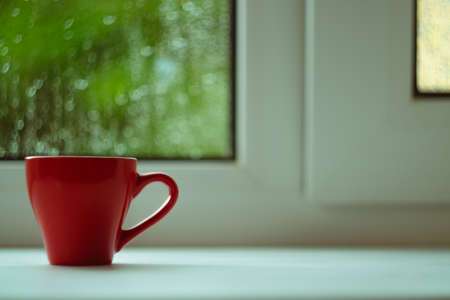 The red espresso cup on white windowsill. On background window and rain. Cup located at left of frame.の写真素材