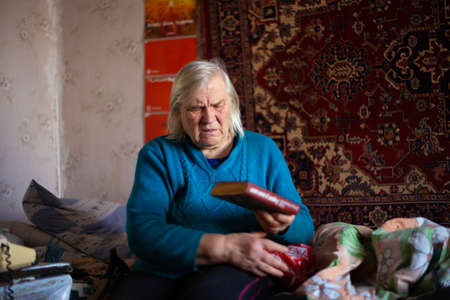 Ukraine, Kyiv, December 12 2018. Old poor gray hair woman holds book in her hand. Woman is sad. Poor life in village. Old age not good. Low-light photo. Editorial Use Only.のeditorial素材