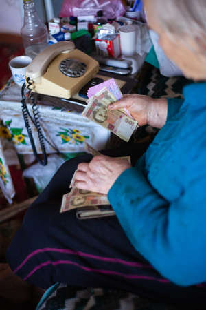 Old poor gray hair woman holds Ukrainian paper money in her hands. Woman is sad. Poor life in village. Old age not good. Low-light photo.の写真素材