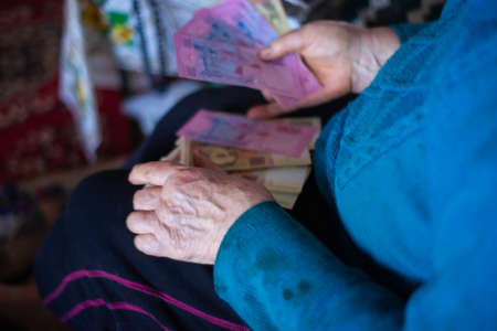 Old poor gray hair woman holds Ukrainian paper money in her hands. Woman is sad. Poor life in village. Old age not good. Low-light photo.の写真素材