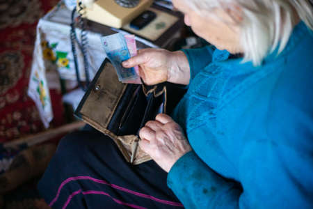 Old poor gray hair woman putting paper Ukrainian money in her vintage leather wallet by her hands. Woman is sad. Poor life in village. Old age not good. Low-light photo.の写真素材