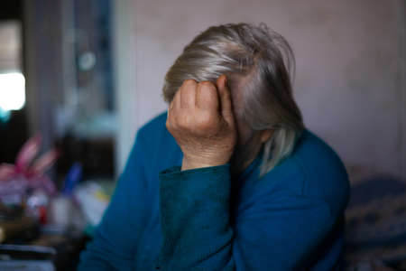 Old poor gray hair woman covered her face with her hand. Woman is sad. Poor life in village. Old age not good. Low-light photo.の写真素材