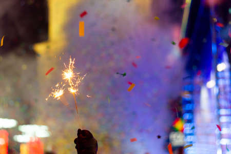 New Year celebration on main square. A lot of smoke or fog, lights, salute and confetti. Blurred colorful background. Bengali fire in hand with glove on foreground. Concert scene with blue light at right side of frame.の写真素材