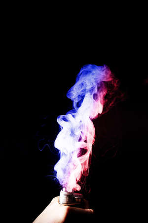 Male hand holds vape. Vape clouds splash from vape coil on black background. Fog is dual color...