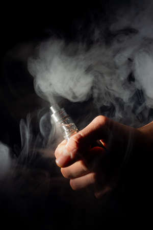 Male hand holds vape in thick vape fog or smoke. Vape clouds around hand with vape on black...