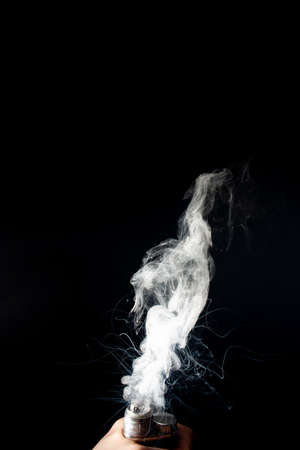 Male hand holds vape without atomiser. Column of vape clouds splash from coil on black background....