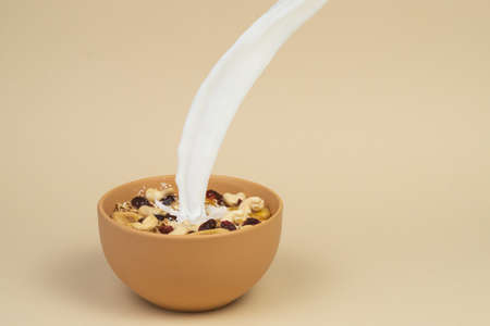 A stream og milk pours onto granola with dried and candied fruits and nuts in beige colour bowl....