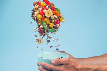 Male hands holding an empty blue bowl on blue background. Candied fruits and nuts flying above the...