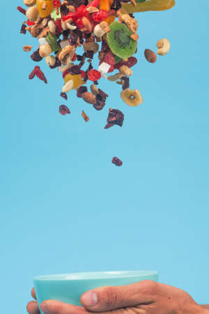 Male hands holding an empty blue bowl on blue background. Candied fruits and nuts flying above the...