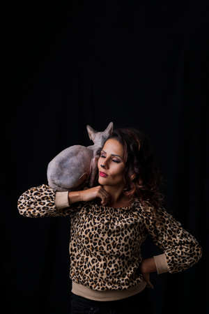 Adorable woman holding a sphinx cat in her arms. Woman in leopard print clothes with cat ears on her head isolated on black background.の写真素材