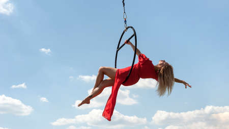 Elegant girl in a red acrobat beautiful dress, performing gymnastic stunts on hoop outdoors against the blue sky with cloudsの写真素材