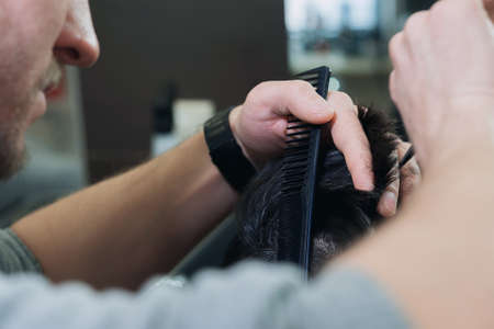 Master hairdresser cutting a short cut young man in a barber shop. Young dark-haired man client in a bright cape sitting on the chair at barbershop.の写真素材