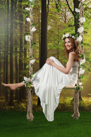Young girl in a long white dress, with a wreath on a swing in the studioの写真素材
