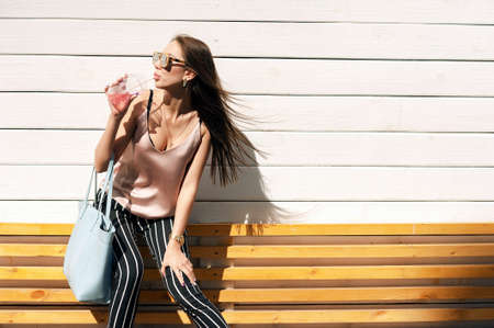 Outdoor portrait of a young beautiful fashionable happy lady wearing sunglasses, in a wooden frame, against a wooden, white wall. The model wears stylish clothes, a beige blouse, black breeches in a white stripe, a bag of light blue. The girl looks away aの写真素材