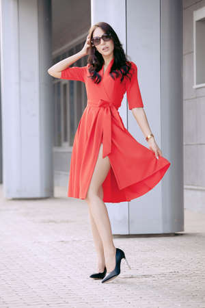 Beautiful brunette young woman in a red dress, in black high-heeled shoes, sunglasses, walking along the street. Fashion photoの写真素材