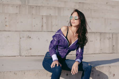 Fashionable young woman in sunglasses, lilac short windbreaker and jeans. Sits on a concrete structure, bare shoulder. Women's fashion. Urban way of life.の写真素材