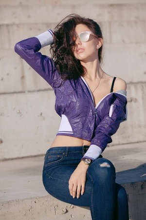 Fashionable young woman in sunglasses, lilac short windbreaker and jeans. Sits on a concrete structure, bare shoulder. Women's fashion. Urban way of life.の写真素材