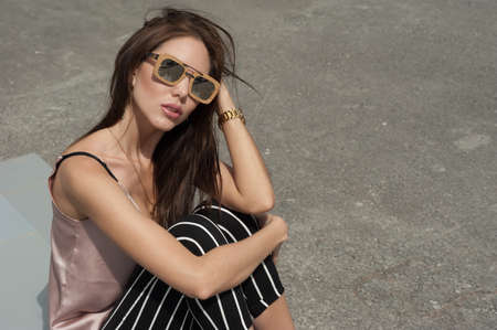 Outdoor portrait of a young beautiful fashionable happy lady wearing sunglasses, in a wooden frame. The model wears stylish clothes, a beige blouse, black breeches in a white stripe, a bag of light blue. The girl is looking at the camera. Women's fashion.の写真素材