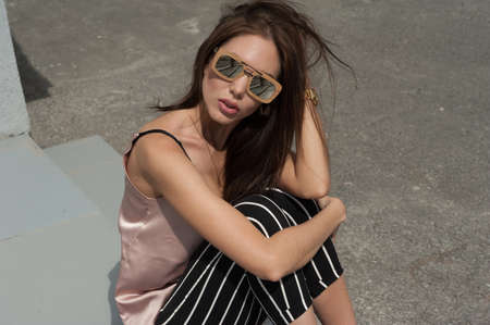 Outdoor portrait of a young beautiful fashionable happy lady wearing sunglasses, in a wooden frame. The model wears stylish clothes, a beige blouse, black breeches in a white stripe, a bag of light blue. The girl is looking at the camera. Women's fashion.の写真素材