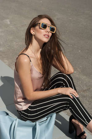 Outdoor portrait of a young beautiful fashionable happy lady wearing sunglasses, in a wooden frame. The model wears stylish clothes, a beige blouse, black breeches in a white stripe, a bag of light blue. The girl is looking at the camera. Women's fashion.の写真素材