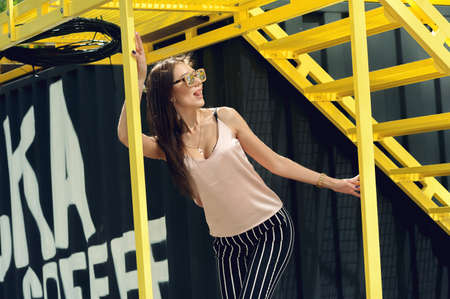 A beautiful girl with sunglasses in a beige T-shirt and black breeches in a white stripe. Against the background of a black container with a yellow staircase. Artistic photography. beauty and fashion, style.の写真素材