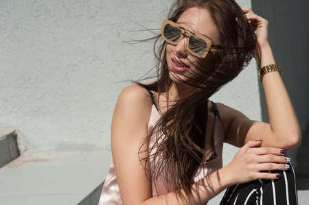 Outdoor portrait of a young beautiful fashionable happy lady, wearing sunglasses, in a wooden frame, creating a city on the street. The model wears stylish clothes. The girl is looking up, the hair is developing in the wind. Women's fashion. Urban way of の写真素材
