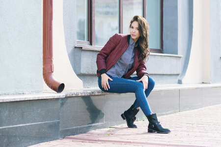 Young beautiful stylish woman in a burgundy pilot's jacket and blue jeans, street style, spring and autumn trend, look, hair in the wind.の写真素材