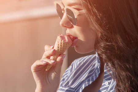 Young, beautiful brunette woman eating pink ice cream in a waffle horn. She wears glasses with dark glasses, and a shirt in a white-blue stripの写真素材