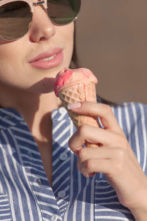 Young, beautiful brunette woman eating pink ice cream in a waffle horn. She wears glasses with dark glasses, and a shirt in a blue and white strip. Close-up photoの写真素材