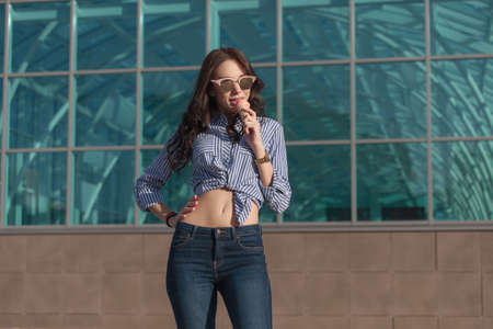 Young, beautiful brunette woman eating a pink ice cream in a waffle horn. She's in a good mood She wears glasses with dark glasses, a shirt in a white and blue strip, and dark blue jeans.の写真素材