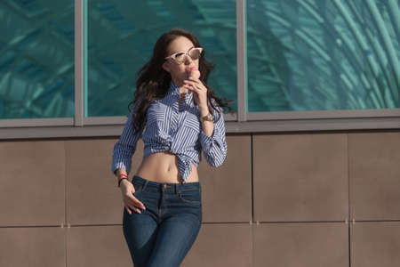 Young, beautiful brunette woman eating a pink ice cream in a waffle horn. She's in a good mood She wears glasses with dark glasses, a shirt in a white and blue strip, and dark blue jeans.の写真素材
