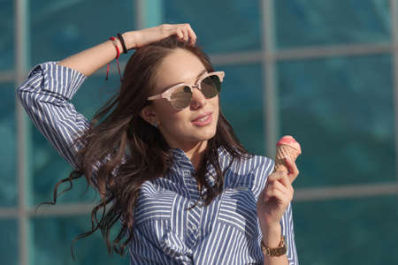 Young, beautiful brunette woman eating pink ice cream in a waffle horn. She's in a good mood She wears glasses with dark glasses, and a shirt in a blue and white strip. Close-up photo.の写真素材
