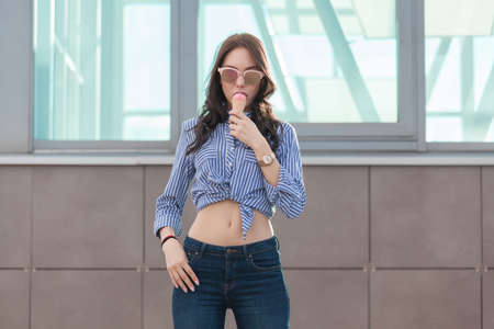 Young, beautiful brunette woman eating a pink ice cream in a waffle horn. She's in a good mood She wears glasses with dark glasses, a shirt in a white and blue strip, and dark blue jeans.の写真素材