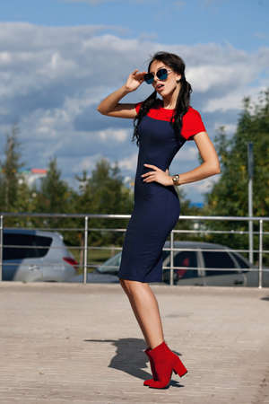 Beautiful brunette young woman in blue and red dress, wearing red high-heeled shoes, sunglasses, walking along the streetの写真素材
