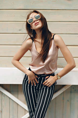 Beautiful woman, wearing dark glasses, against the background of a tree wall. The model wears stylish clothes, beige knitwear and black breeches in a white stripe. Hair develops in the wind. Women's fashion. Urban lifestyle.の写真素材