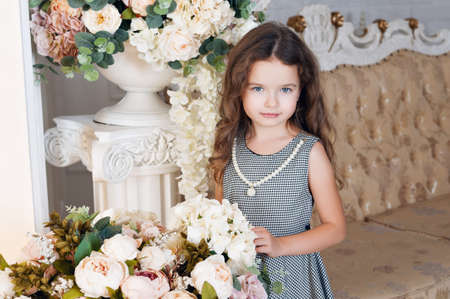A little girl of seven with chestnut hair, stands next to a bouquet of peony flowers, smiling, in a gray dress in a cage, curls. The concept of a holiday.の写真素材