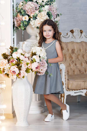 A little girl of seven with chestnut hair, stands next to a bouquet of peony flowers, smiling, in a gray dress in a cage, curls. The concept of a holiday.の写真素材