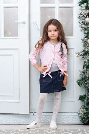 A nice little girl of seven years with chestnut-colored hair, stands on the background of the folding doors decorated in a Christmas style, in a blue skirt, a pink jacket, posing for the camera. Concept of a holiday, Christmas.の写真素材