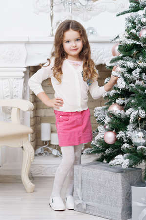 A nice little girl of seven with chestnut curly hair, with a Christmas tree and gifts, stands in a pink skirt, a white sweater, posing for the camera. Concept of a holiday, Christmas.の写真素材