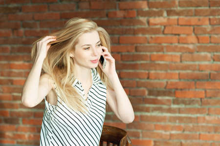 A blonde girl is calling by the phone against a brick wall backgroundの写真素材