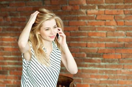A blonde girl is calling by the phone against a brick wall background, calling the apartment repair service. Advertising of construction servicesの写真素材