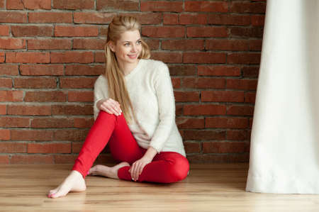 Beautiful young blonde woman in a white warm jacket and red zhdinsah, sits on the floor near the window, against a red brick wallの写真素材