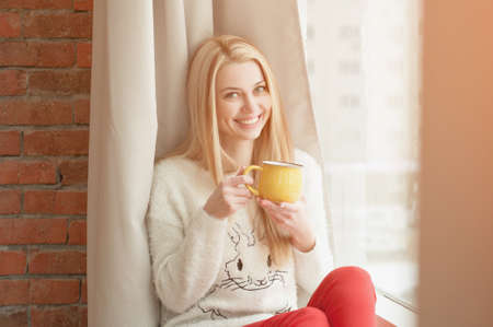 Young woman standing by the window with a cup of coffee, looking outside and smiling. Natural outdoor lightの写真素材