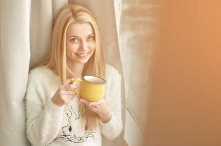Young woman standing by the window with a cup of coffee, looking outside and smiling. Natural outdoor lightの写真素材