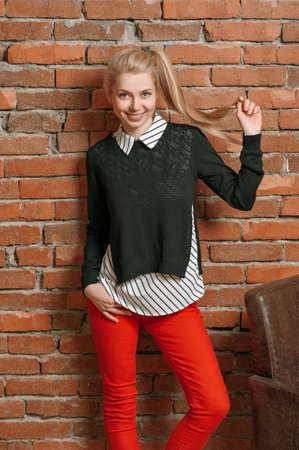 Beautiful young blonde in a white blouse and dark jacket and red jeans. Standing on against a red brick wall, next to a brown leather armchair and holding his own hairの写真素材