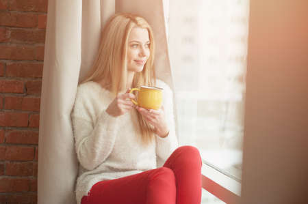 Young woman standing by the window with a cup of coffee, looking outside and smiling. Natural outdoor lightの写真素材