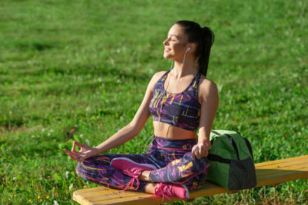 Concept of sport and lifestyle - young woman resting after sports in lotus position, smiling, sitting on bench, outdoors.の写真素材