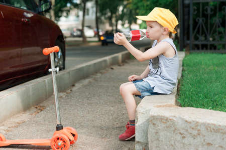 Boy 5 years old, with a scooter on the road in the park. The child drinks water. Family holiday