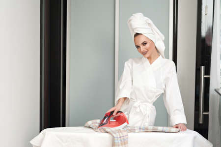 A young cheerful woman after a shower, wearing a white bathrobe and a towel on her head ironing clothes in the home interiorの写真素材